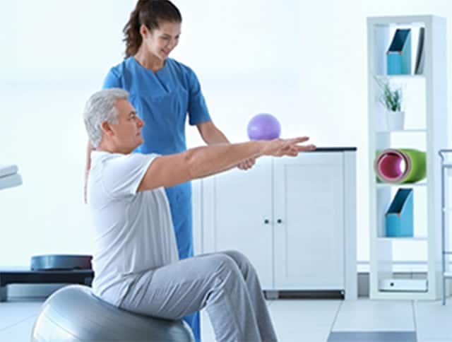 A physiotherapist helping patient to do exercise on fitness ball in physio room.